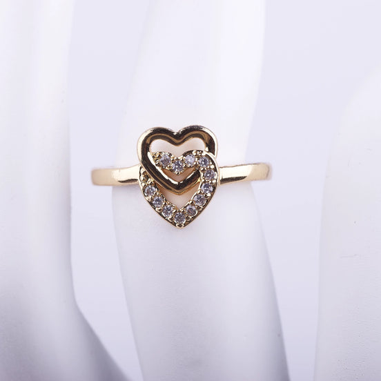 Anillo dorado delgado con dos corazones entrelazados en el centro, uno liso y otro con cristales brillantes sobre fondo blanco.