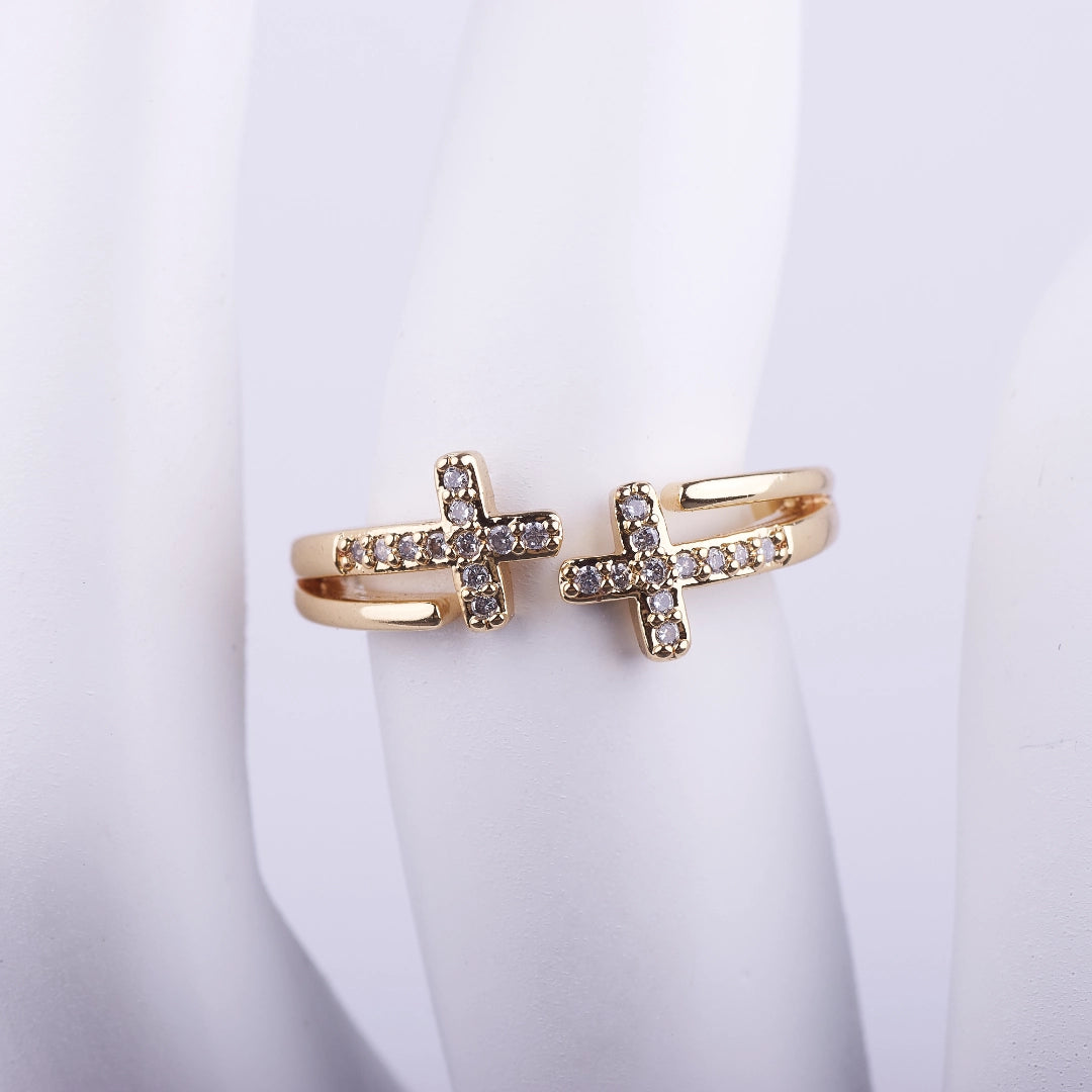 Anillo abierto dorado de doble banda con dos cruces frontales decoradas con cristales brillantes sobre fondo blanco.