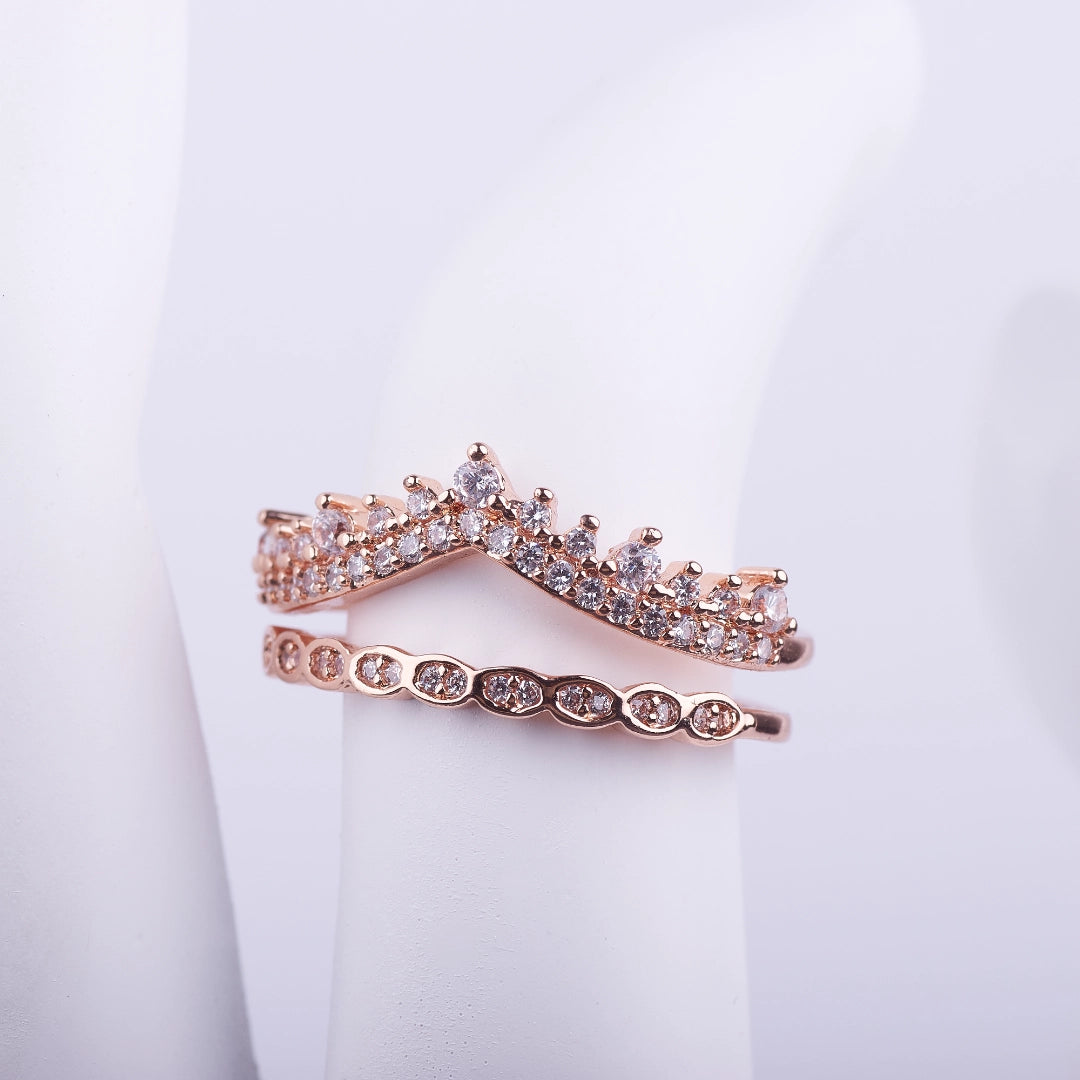 Anillo único de doble banda en oro rosa con diseño de corona y alianza de cristales unidas por la base sobre fondo blanco.