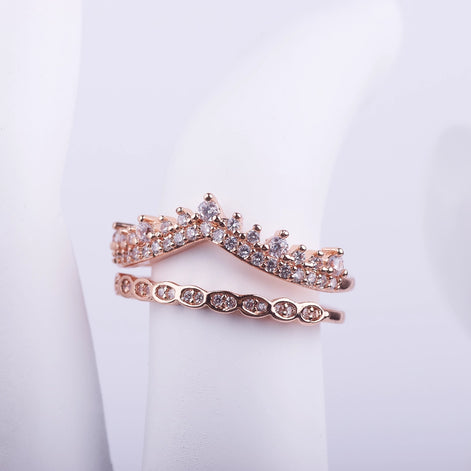 Anillo único de doble banda en oro rosa con diseño de corona y alianza de cristales unidas por la base sobre fondo blanco.