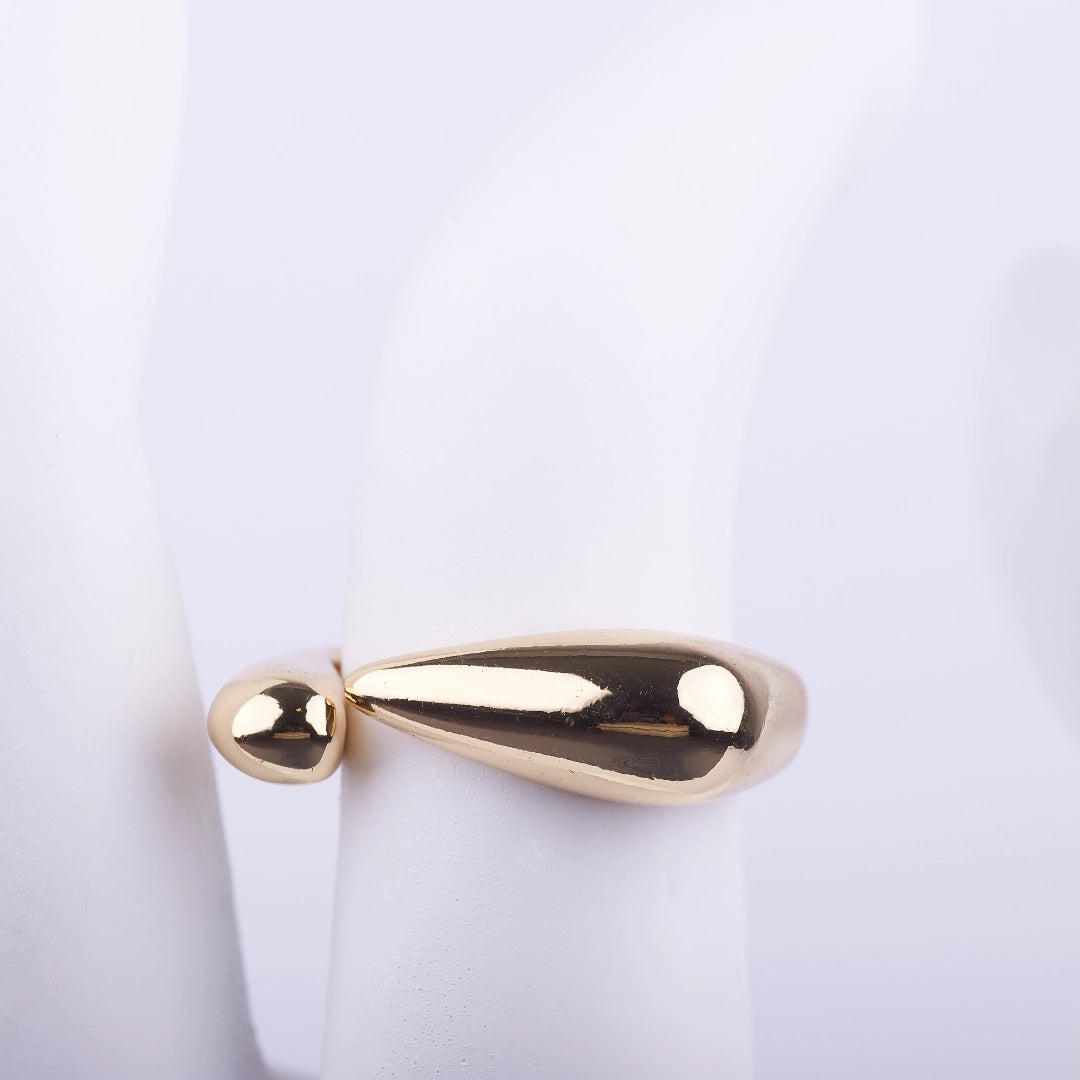 Anillo abierto dorado con extremos en forma de gota fluida y acabado brillante sobre fondo blanco.