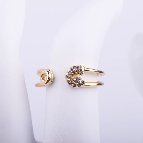 Anillo abierto con forma de imperdible dorado y detalles de cristales brillantes en la punta, sobre fondo blanco.