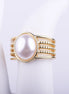 Anillo ancho dorado con una gran perla blanca central y banda de múltiples filas lisas y de cuentas sobre fondo blanco