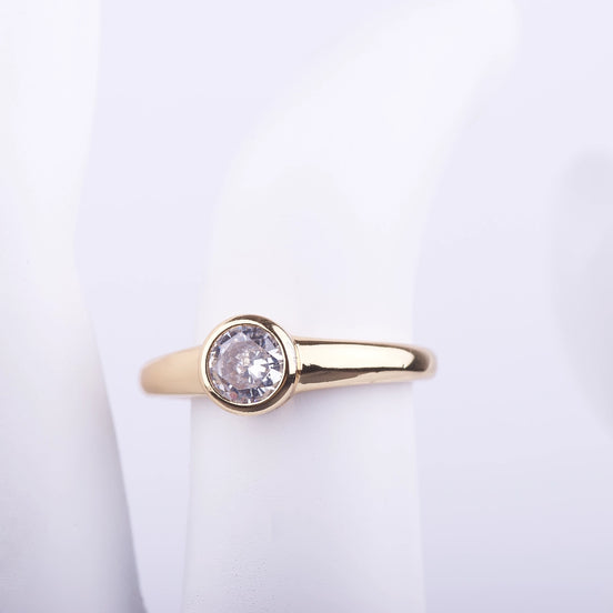 Anillo dorado con cristal central redondo protegido por un marco de metal circular liso sobre fondo blanco.