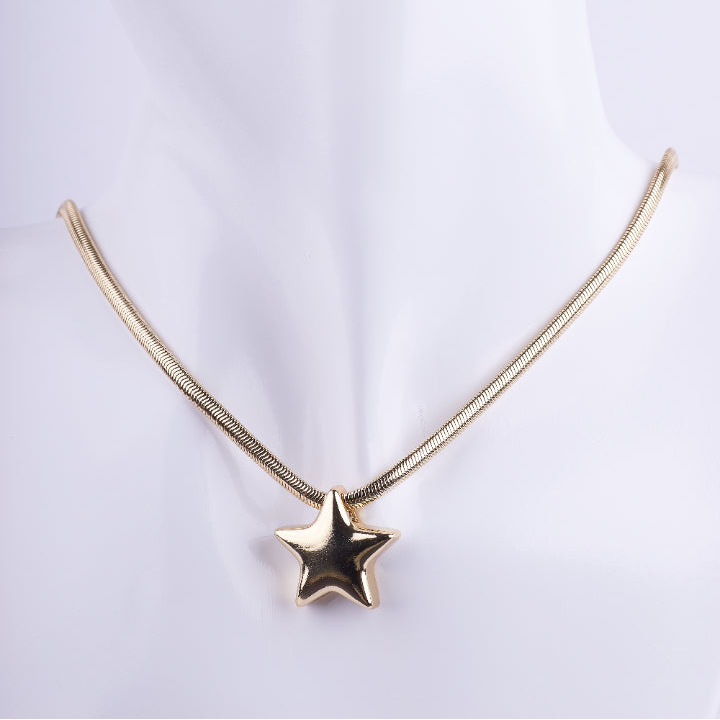 Collar estilo choker de cadena serpiente (snake chain) dorada con un dije de estrella voluminoso (puffy) y liso. El dije tiene un acabado de metal espejo brillante, exhibido en un busto de maniquí blanco.