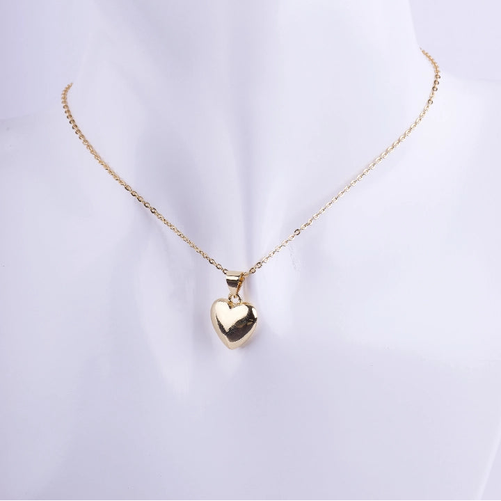 Collar de cadena de eslabones dorados finos con un dije de corazón inflado (puffy) de metal liso y muy pulido. El corazón es de color dorado y está exhibido en un busto de maniquí blanco.
