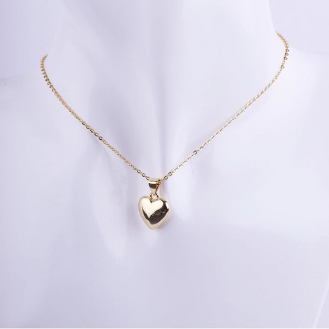 Collar de cadena de eslabones dorados finos con un dije de corazón inflado (puffy) de metal liso y muy pulido. El corazón es de color dorado y está exhibido en un busto de maniquí blanco.