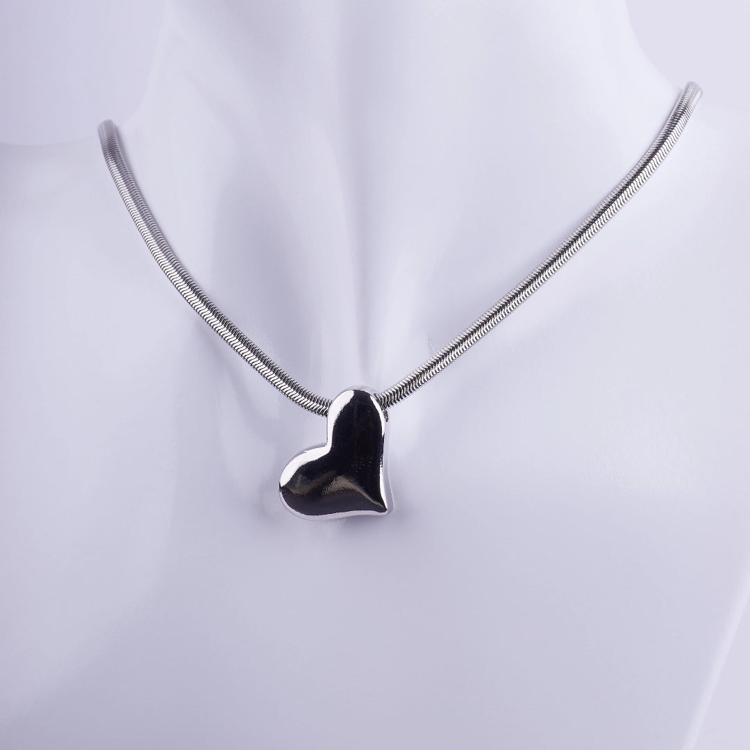Collar de corazón plateado con acabado espejo y cadena tipo serpiente fina sobre un busto de exhibición blanco.