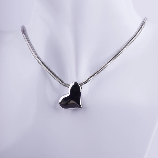 Collar de corazón plateado con acabado espejo y cadena tipo serpiente fina sobre un busto de exhibición blanco.