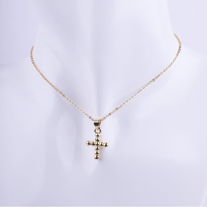 Collar de cadena de eslabones finos dorados con un dije de cruz tridimensional formado por pequeñas esferas doradas en relieve. Exhibido en un busto de maniquí blanco.