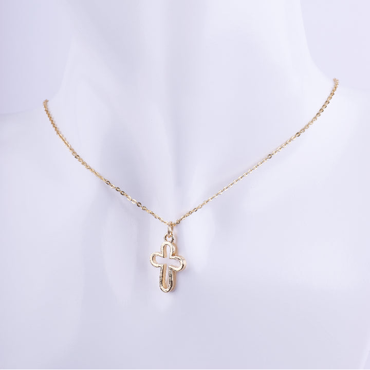 Collar de cadena de eslabones dorados finos con un dije de cruz con diseño calado en el centro. El dije es de metal pulido y brillante, exhibido en un busto de maniquí blanco.