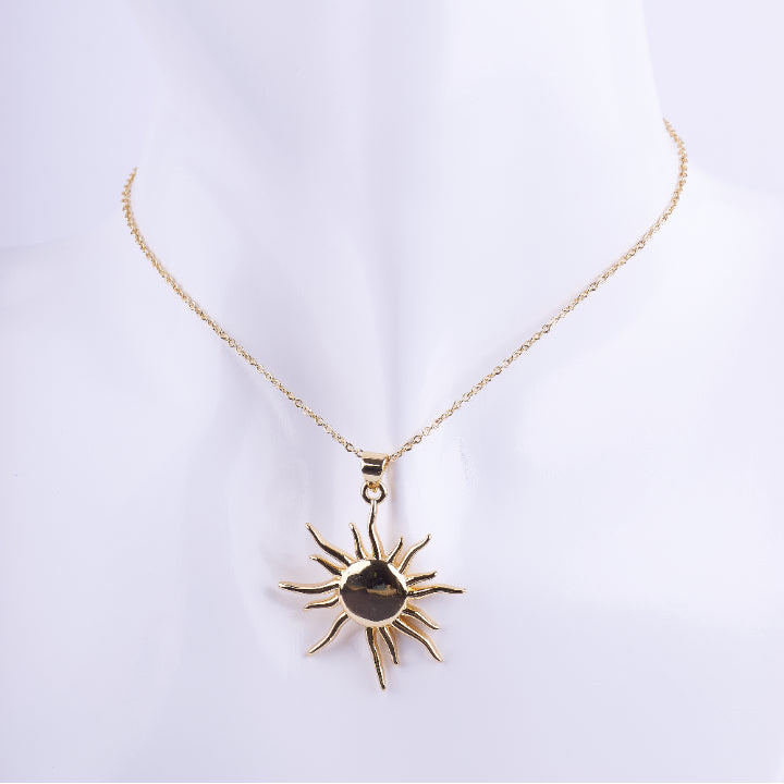 Collar de cadena de eslabones dorados finos con un dije grande de sol con rayos puntiagudos. exhibido en un busto de maniquí blanco.