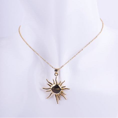 Collar de cadena de eslabones dorados finos con un dije grande de sol con rayos puntiagudos. exhibido en un busto de maniquí blanco.