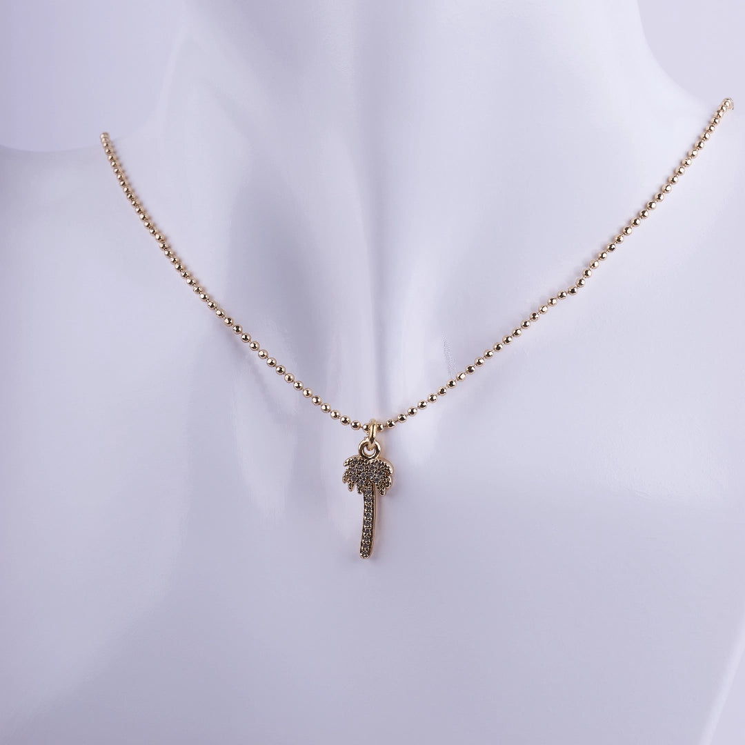 Collar dorado con dije de palmera brillante y cadena de esferas pequeñas sobre busto blanco, joyería de estilo tropical y elegante.
