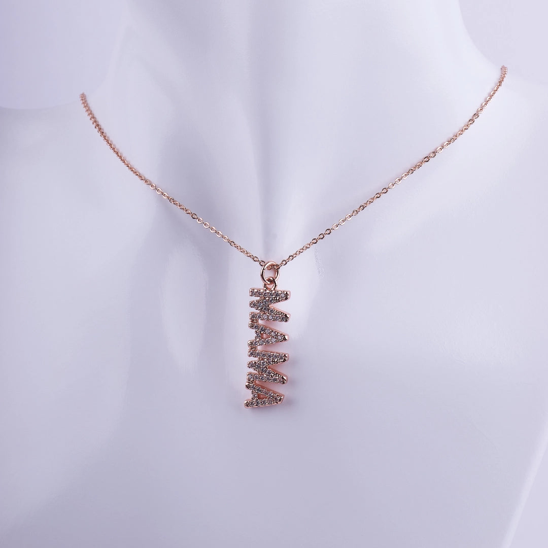 Collar con dije vertical que dice 'MAMA' adornado con zirconias brillantes en baño de oro rosa sobre un fondo neutro.