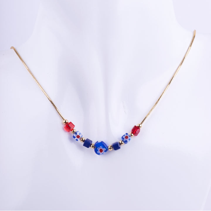 Collar de cadenilla dorada con nueve cuentas de cristal de Murano de colores rojo y azul, exhibido en un busto blanco. El detalle central es una cuenta azul con diseño de flor roja y blanca.