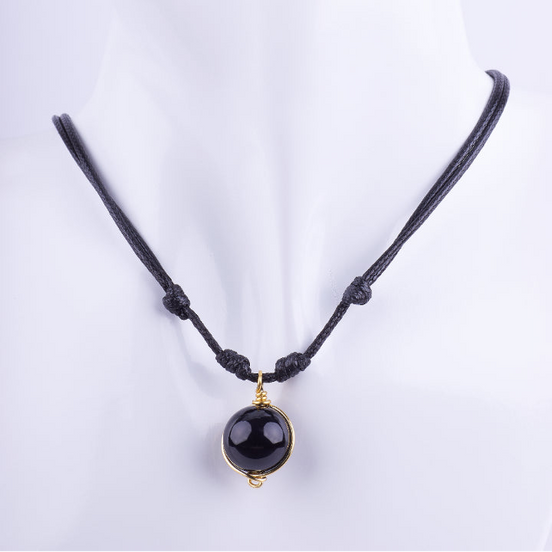 Collar estilo choker de cordón doble de macramé negro con un dije de esfera de piedra negra (Obsidiana u Ónix) envuelto en alambre dorado. El cordón es ajustable con nudos, exhibido en un busto de maniquí blanco.