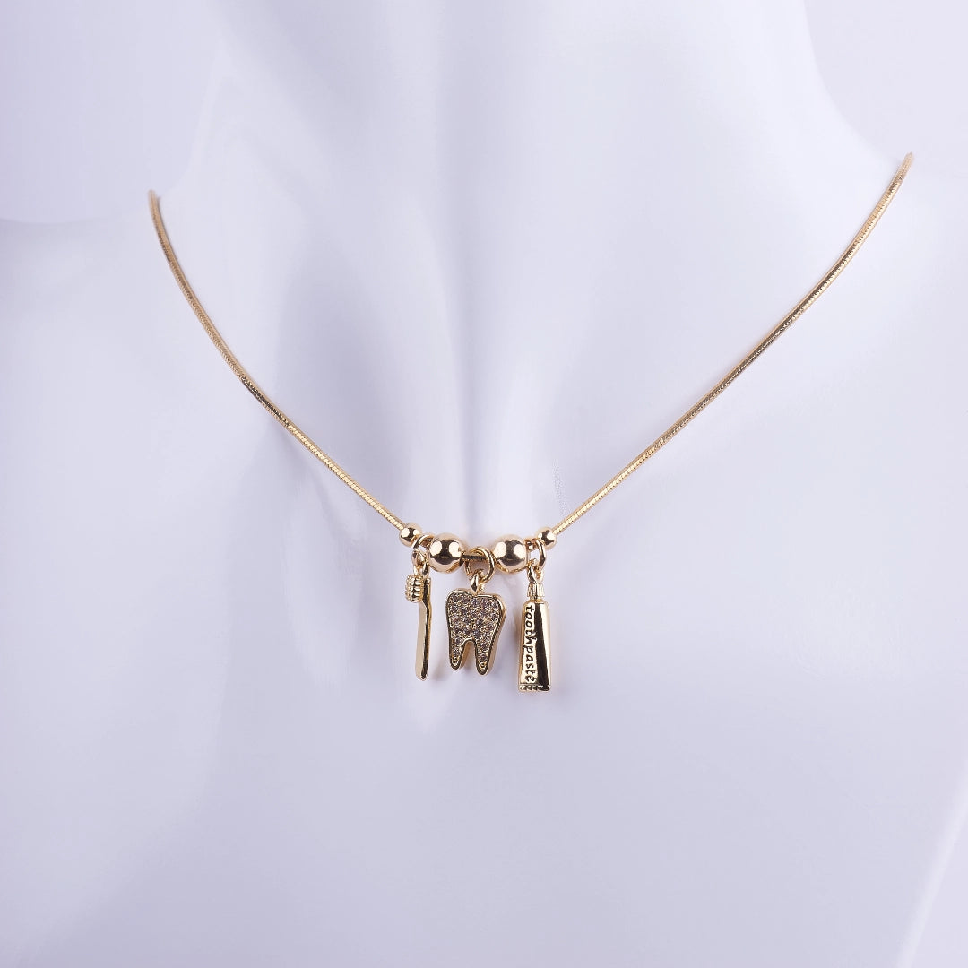 Collar de odontología bañado en oro con tres dijes: una muela con zirconias, un cepillo de dientes y una pasta dental sobre cadena de serpiente.