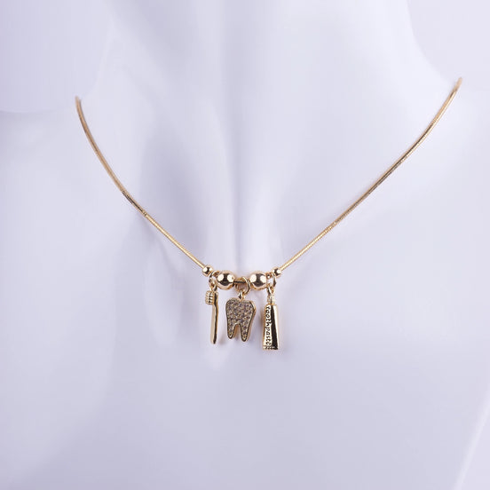 Collar de odontología bañado en oro con tres dijes: una muela con zirconias, un cepillo de dientes y una pasta dental sobre cadena de serpiente.