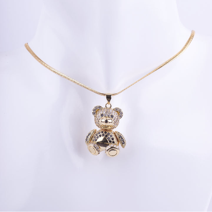 Collar de cadenilla fina dorada con un dije tridimensional de un osito de peluche sentado. El osito está cubierto de cristales brillantes y tiene detalles en negro, exhibido en un busto de maniquí blanco