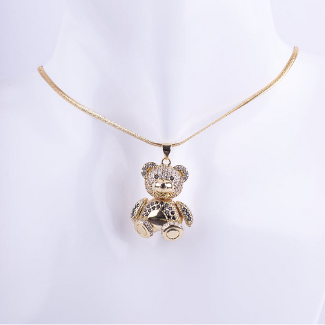 Collar de cadenilla fina dorada con un dije tridimensional de un osito de peluche sentado. El osito está cubierto de cristales brillantes y tiene detalles en negro, exhibido en un busto de maniquí blanco