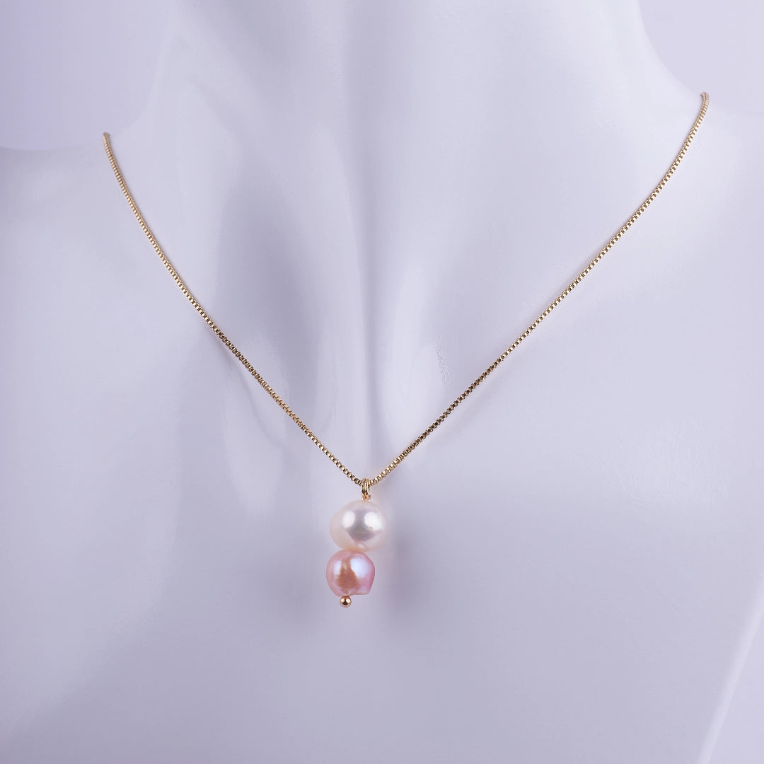 Collar dorado con dos perlas naturales colgando verticalmente, una blanca y una rosa, sobre una cadena veneciana fina.