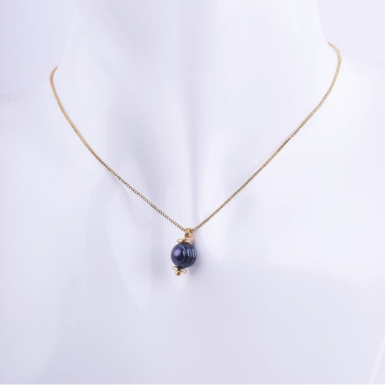 Collar de cadena tipo veneciana dorada con un dije de una perla negra redonda. La perla está coronada por una pequeña cuenta dorada, exhibida en un busto de maniquí blanco.