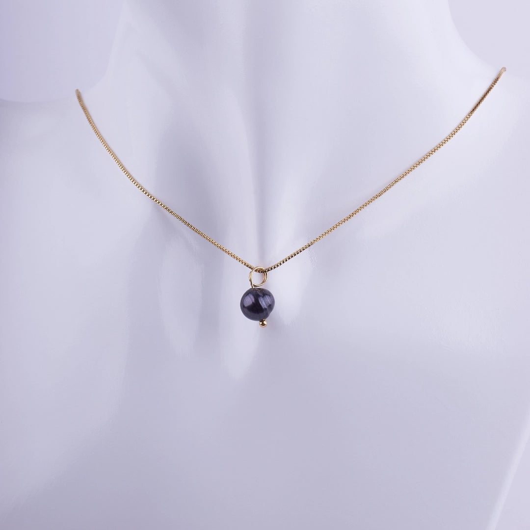 Collar minimalista con una perla negra solitaria colgando de una cadena veneciana bañada en oro sobre un fondo blanco.