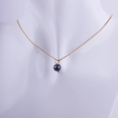 Collar minimalista con una perla negra solitaria colgando de una cadena veneciana bañada en oro sobre un fondo blanco.