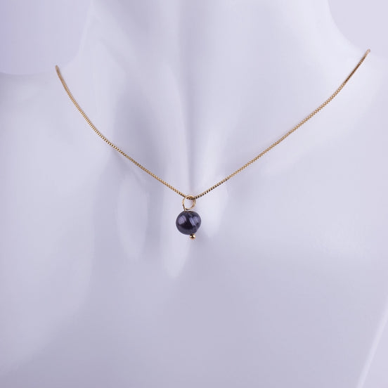 Collar minimalista con una perla negra solitaria colgando de una cadena veneciana bañada en oro sobre un fondo blanco.