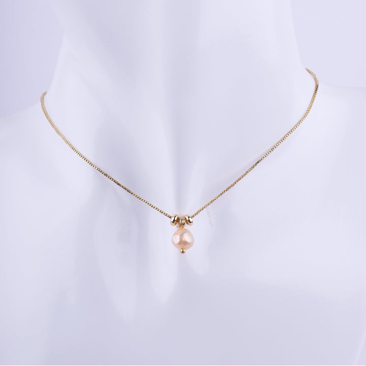 Collar de cadena de eslabones dorados finos con un dije de una perla natural redonda en tono rosado melocotón, flanqueada por dos cuentas doradas. Exhibido en un busto de maniquí blanco.