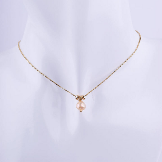 Collar de cadena de eslabones dorados finos con un dije de una perla natural redonda en tono rosado melocotón, flanqueada por dos cuentas doradas. Exhibido en un busto de maniquí blanco.