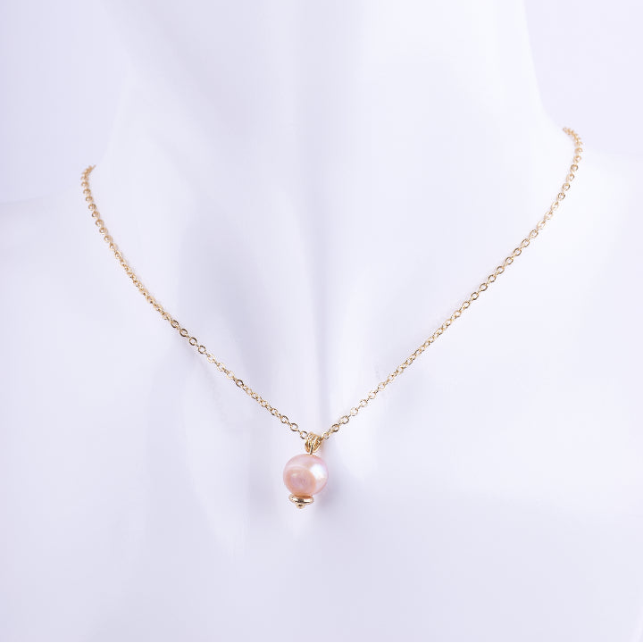 Collar de cadena de eslabones dorados finos con un dije de una perla natural redonda en tono rosado o melocotón. La perla está coronada por una cuenta dorada, exhibido en un busto de maniquí blanco.