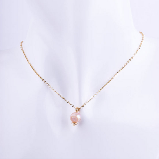Collar de cadena de eslabones dorados finos con un dije de una perla natural redonda en tono rosado o melocotón. La perla está coronada por una cuenta dorada, exhibido en un busto de maniquí blanco.