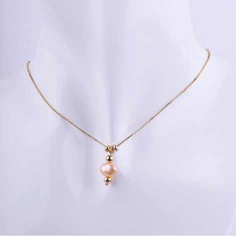 Collar de cadena veneciana dorada con un dije de perla natural en forma ovalada o alargada de color rosado melocotón, flanqueada por dos esferas doradas. Exhibido en un busto de maniquí blanco.