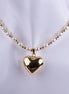 Collar de perlas blancas y esferas doradas con un gran dije de corazón pulido en baño de oro sobre un fondo neutro.