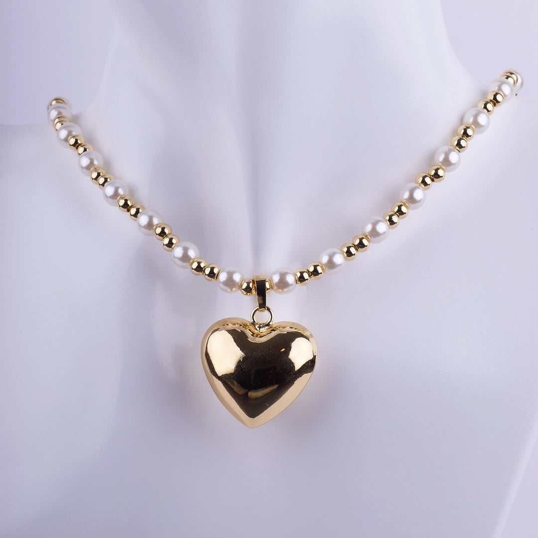 Collar de perlas blancas y esferas doradas con un gran dije de corazón pulido en baño de oro sobre un fondo neutro.