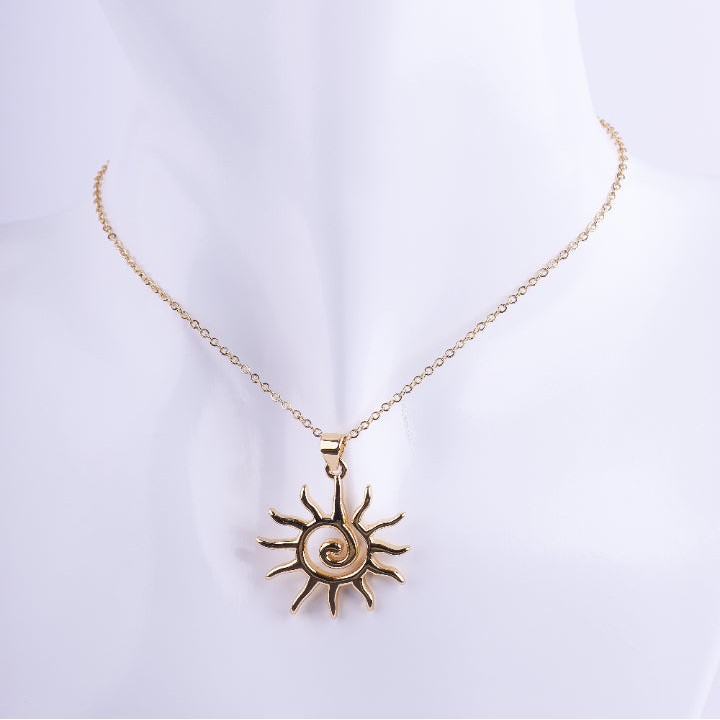 Collar de cadena de eslabones dorados finos con un dije grande de sol con rayos y una espiral pulida en el centro. El dije es de metal pulido y brillante, exhibido en un busto de maniquí blanco