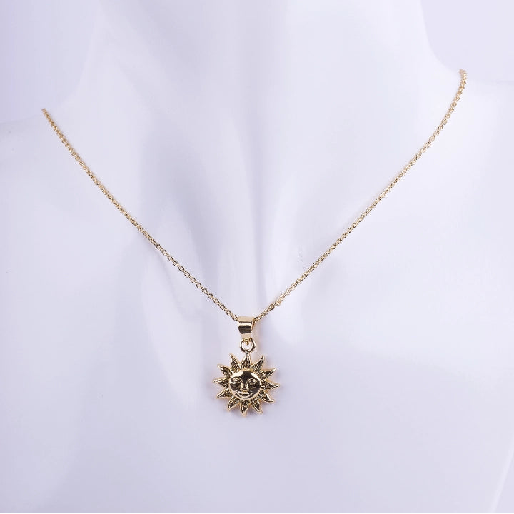 Collar de cadena de eslabones finos dorados con un dije de sol con rayos y un rostro central. El dije tiene un acabado de metal liso brillante, exhibido en un busto de maniquí blanco.