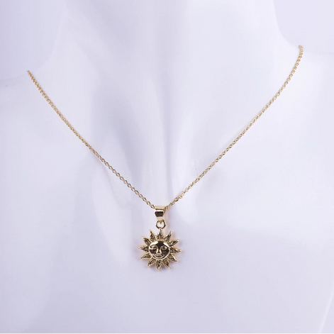 Collar de cadena de eslabones finos dorados con un dije de sol con rayos y un rostro central. El dije tiene un acabado de metal liso brillante, exhibido en un busto de maniquí blanco.