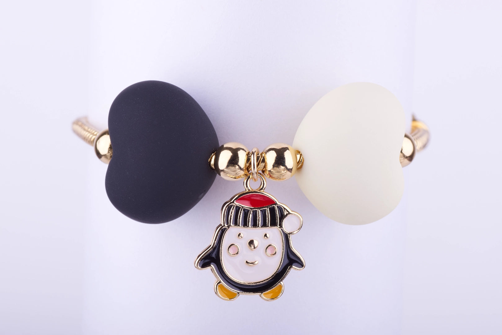 Pulsera dorada con dije de pingüino navideño esmaltado en blanco, negro y rojo, y dos cuentas grandes en forma de corazón, una blanca y otra negra.