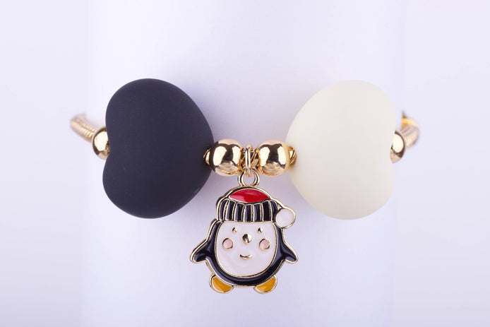 Pulsera dorada con dije de pingüino navideño esmaltado en blanco, negro y rojo, y dos cuentas grandes en forma de corazón, una blanca y otra negra.
