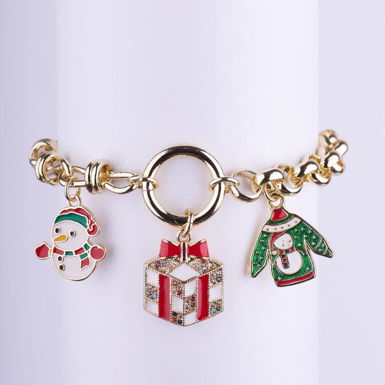 Pulsera chapada en oro de eslabones gruesos con cierre de aro central y tres dijes: un muñeco de nieve, una caja de regalo roja con gemas y un jersey navideño (ugly sweater) verde.